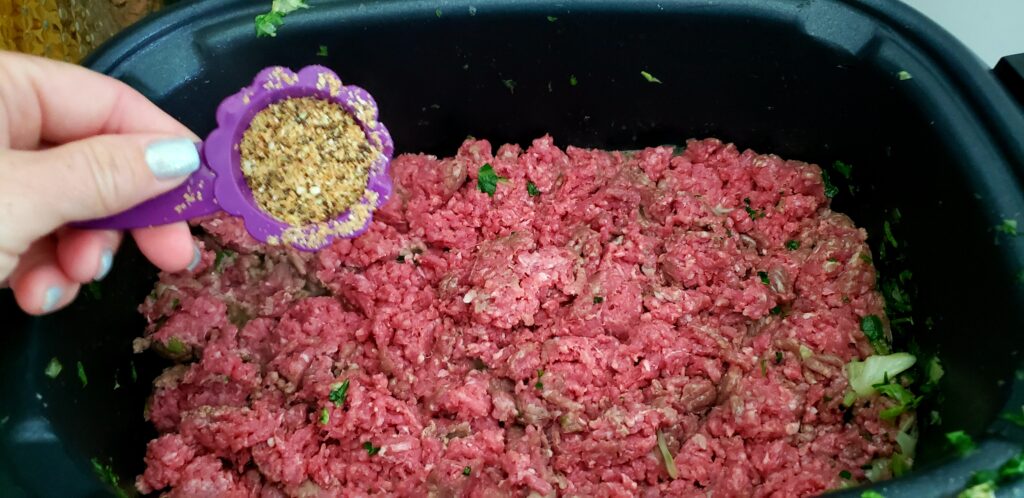 raw ground beef in a crockpot with a hand pouring seeds into the meat