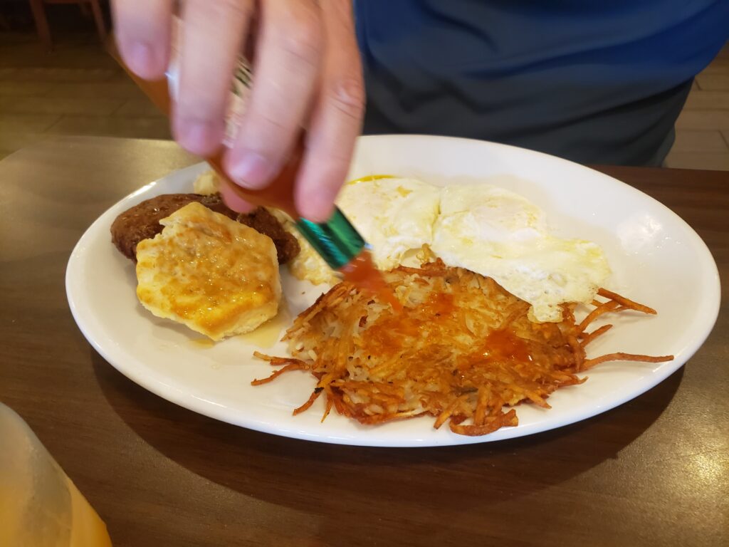 white plate with breakfast food mans hand pouring hot sauce on it